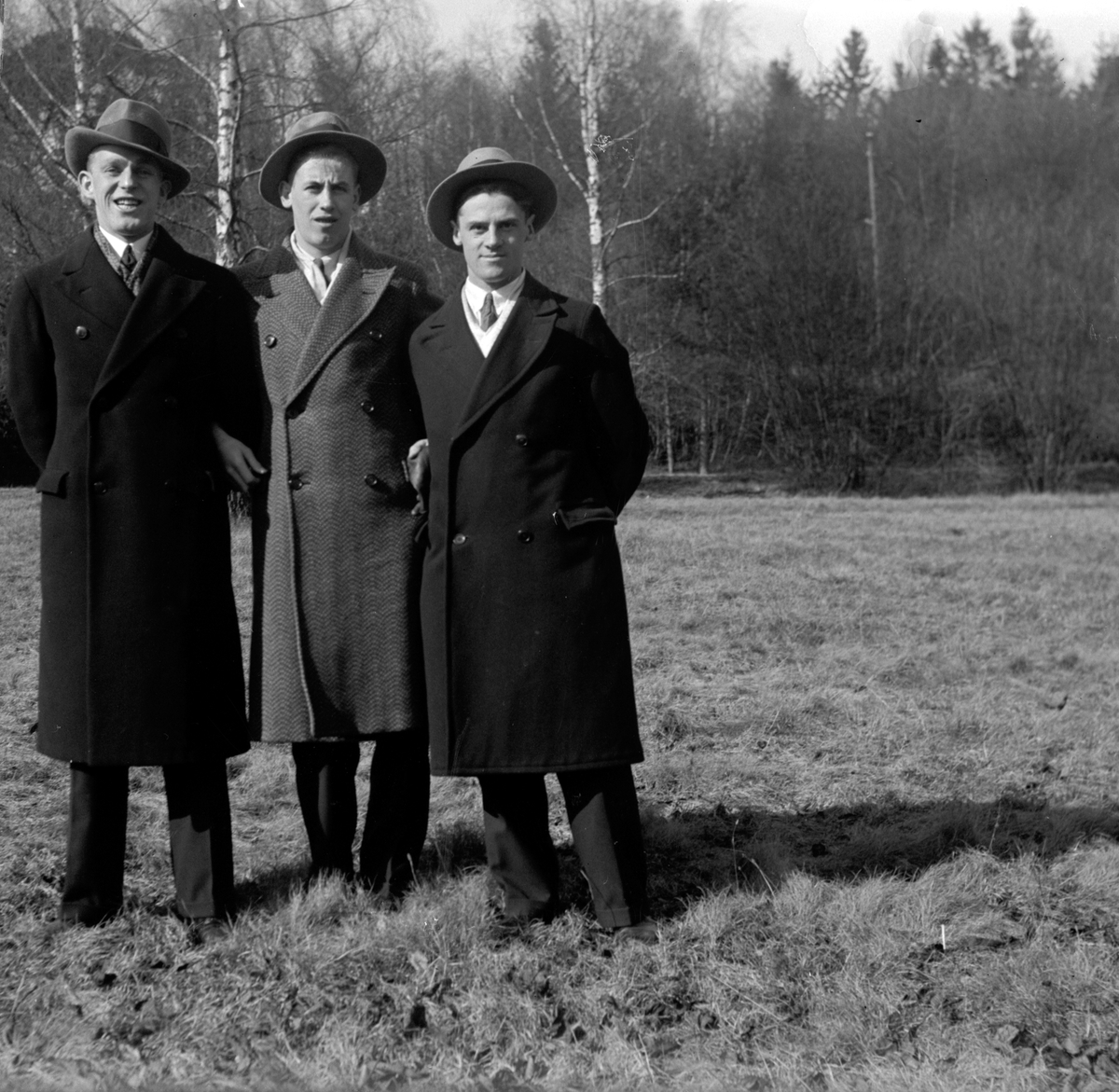Tre män klädda i rock och hatt, står på ett fält med avlövade träd i bakgrunden.