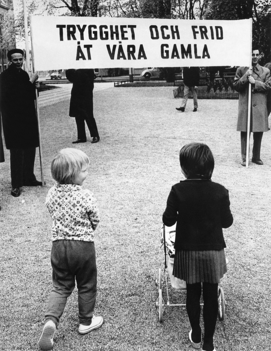 En demonstration med text på banderollen: Trygghet och frid åt våra gamla. Två barn i förgrunden, den ena kör en dockvagn.
