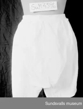 Vita benkläder med knäppning i sidorna med sydd knapp. Benen har 13 cm långa sprund. Hålsöm. Vidhängande lapp med texten 'Märta Moberg'.