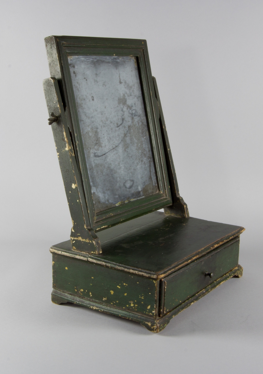 Toalettspegel av trä, grönmålad, med kvadratisk spegel löst monterad med muttrar vid två raka ståndare och med låda med knopp.