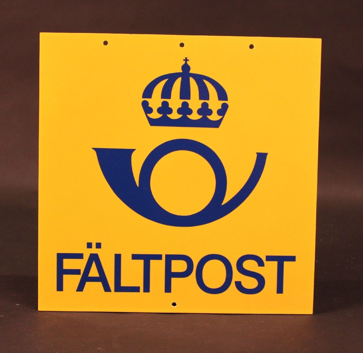 Skylt i papp med gul bakgrund och en blå postsymbol, modell 1965 och ordet: Fältpost i blått i nederkant. Postsymbolen är i plast och är påklistrade.