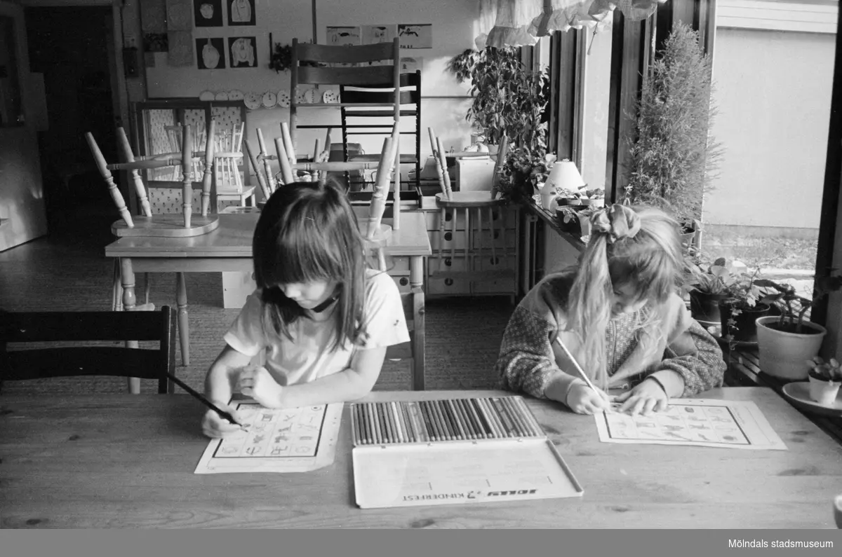 Cecilia och Jessika sitter vid ett bord och färglägger med hjälp av färgpennor, förtryckta figurer på var sitt A4-blad. Båda är mycket koncentrerade. Lunkentussen, Katrinebergs daghem 1992.