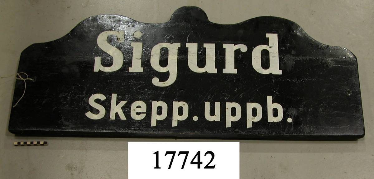 Rekangulär skylt, ovansidan sågad i tre bågar, svart målad. Vit schablonmålad text: " Sigurd Skepp. uppb. ", svartmålad baksida.
