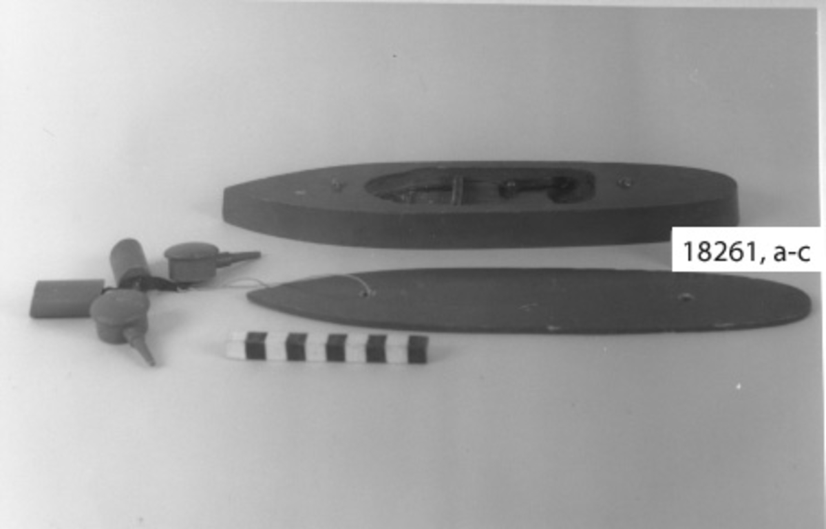 Leksaksbåt av trä i form av pansarbåt målad i grått. Bestående av tre olika delar a: skrov :b däck, :c skorstenar och artilleripjäser.

:a skrov H= 23 mm, L= 298 mm, B=72 mm

Skrovet i ett stycke med spetsig för, vinklad utåt vid vattenlinjen och rundad akter. Plan botten. Invändigt urholkning med mekanism liknande råttfälla. En spiralfjäder spänner en bygel som vid tryck på en klapp placerad på båtern utsida midskepps utlöses. Röd knapp, runt denna flertal hål som visar var projektilerna missat. Vid träff på knappen flyger däck, artilleripjäser och skorstenar iväg!