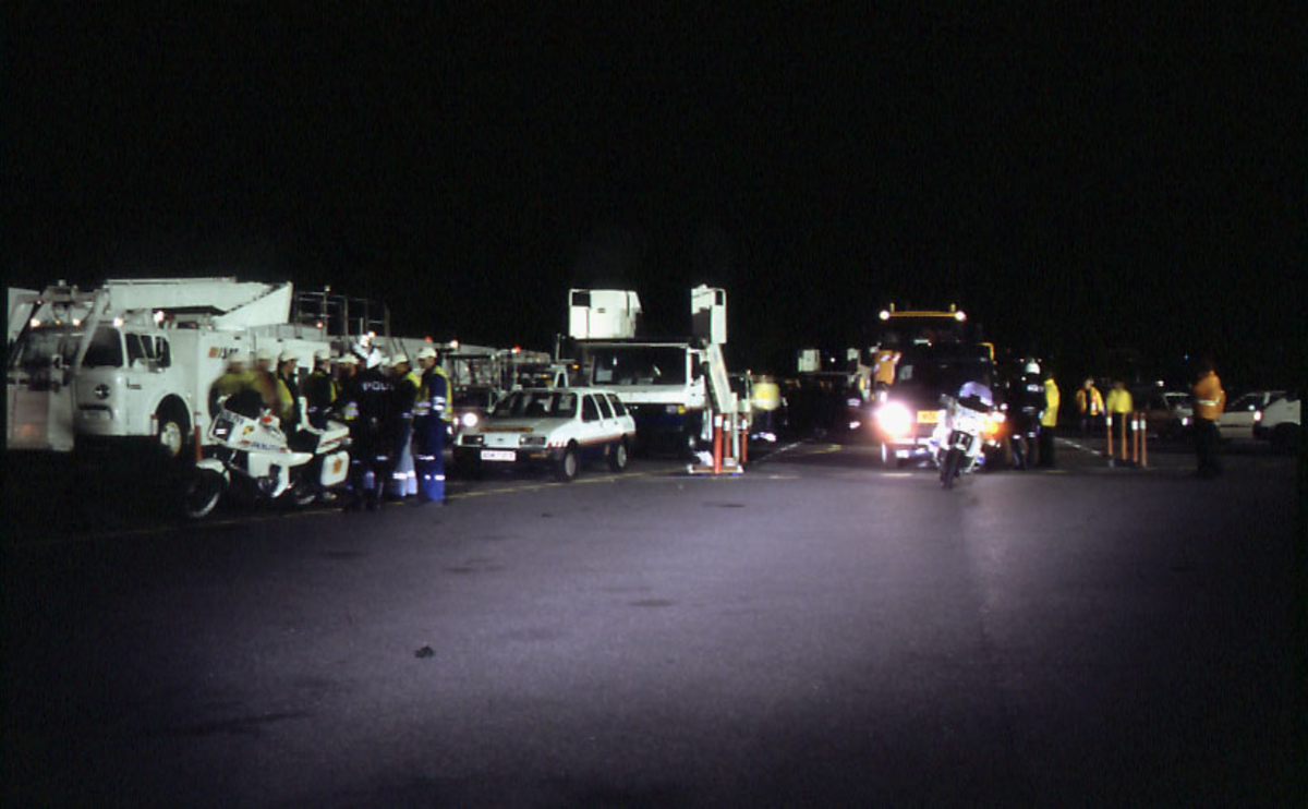 Lufthavn, flere personer, personell og politi, står foran flere kjøretøyer på en del av rullebanen.  Flere personer i bakgrunnen.
