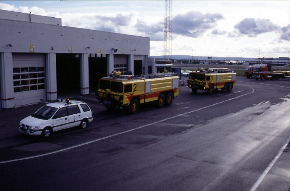 Lufthavn, 3 kjøretøyer fra Luftfartsverket, 2 av dem brannbiler, parkert foran 1 garasjebygning.  Flere kjøretøyer og flyplassbygninger i bakgrunnen.