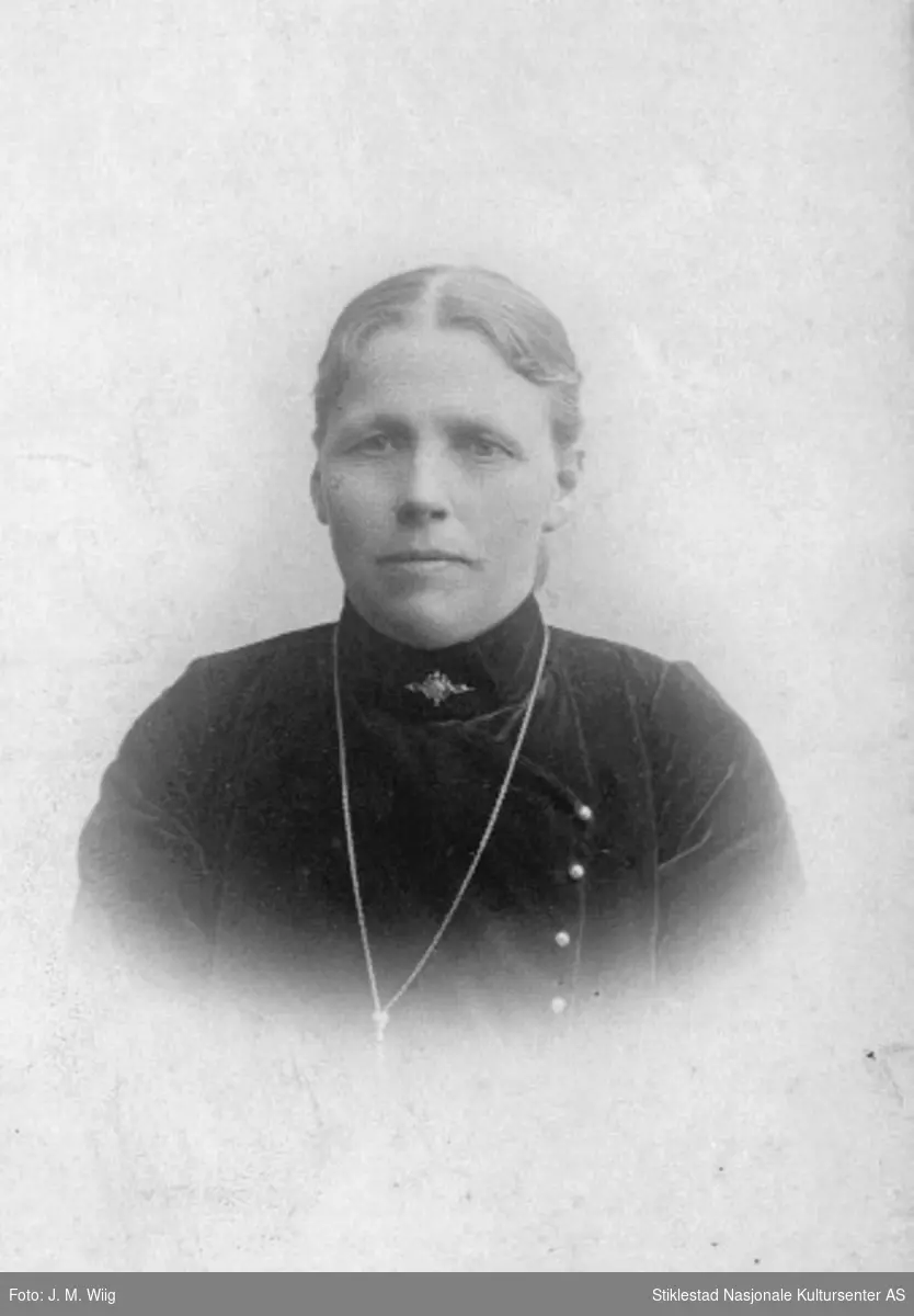 Portrett av ukjent eldre kvinne i mørk høyhalset kjole, smykkelenke og brosje i hals.