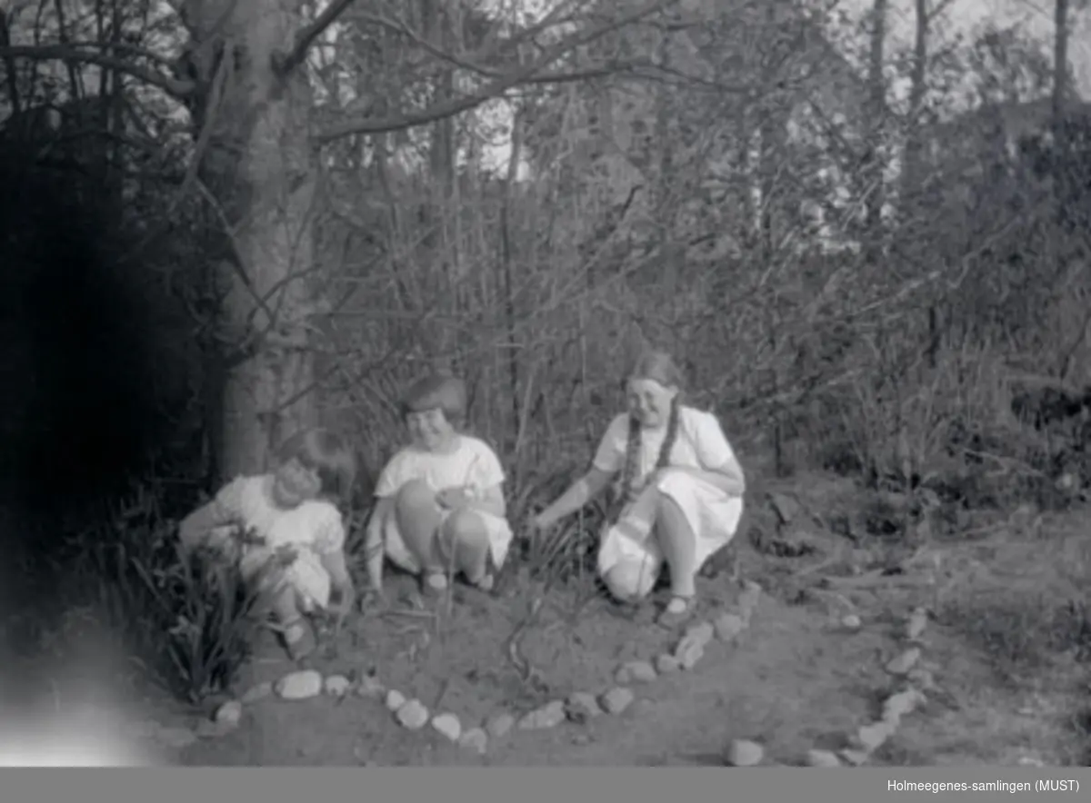 Tre jenter sitter i en skog eller hage ved en steinkantet sti eller blomsterbed.