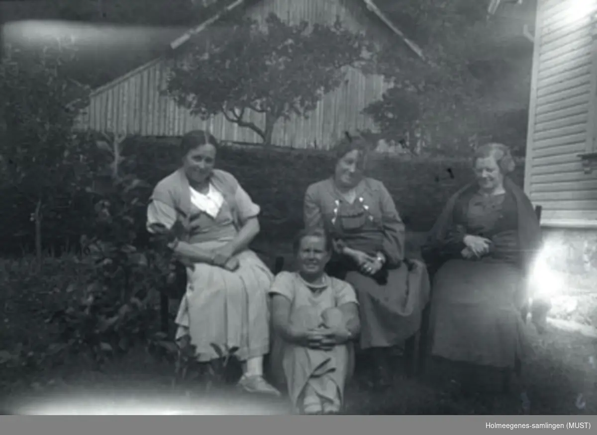 Fire kvinner sitter utendørs. Hjørnet av et hus ses i høyre billedkant, og i bakgrunnen ses gavlen av en driftsbygning.