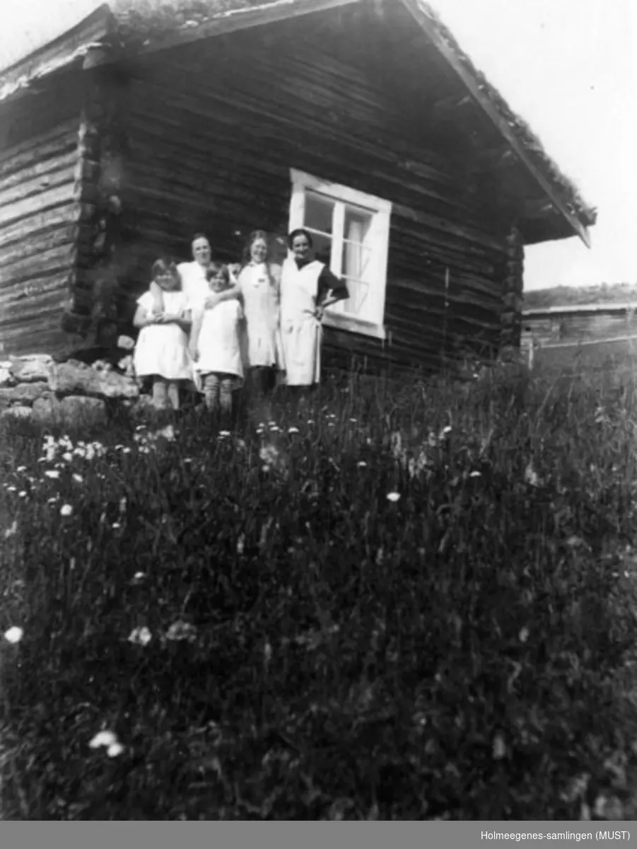 To kvinner og tre jenter står foran en liten tømmerstue med torvtak. Se også ST.K.HE 2007-011-0111 til -0126.