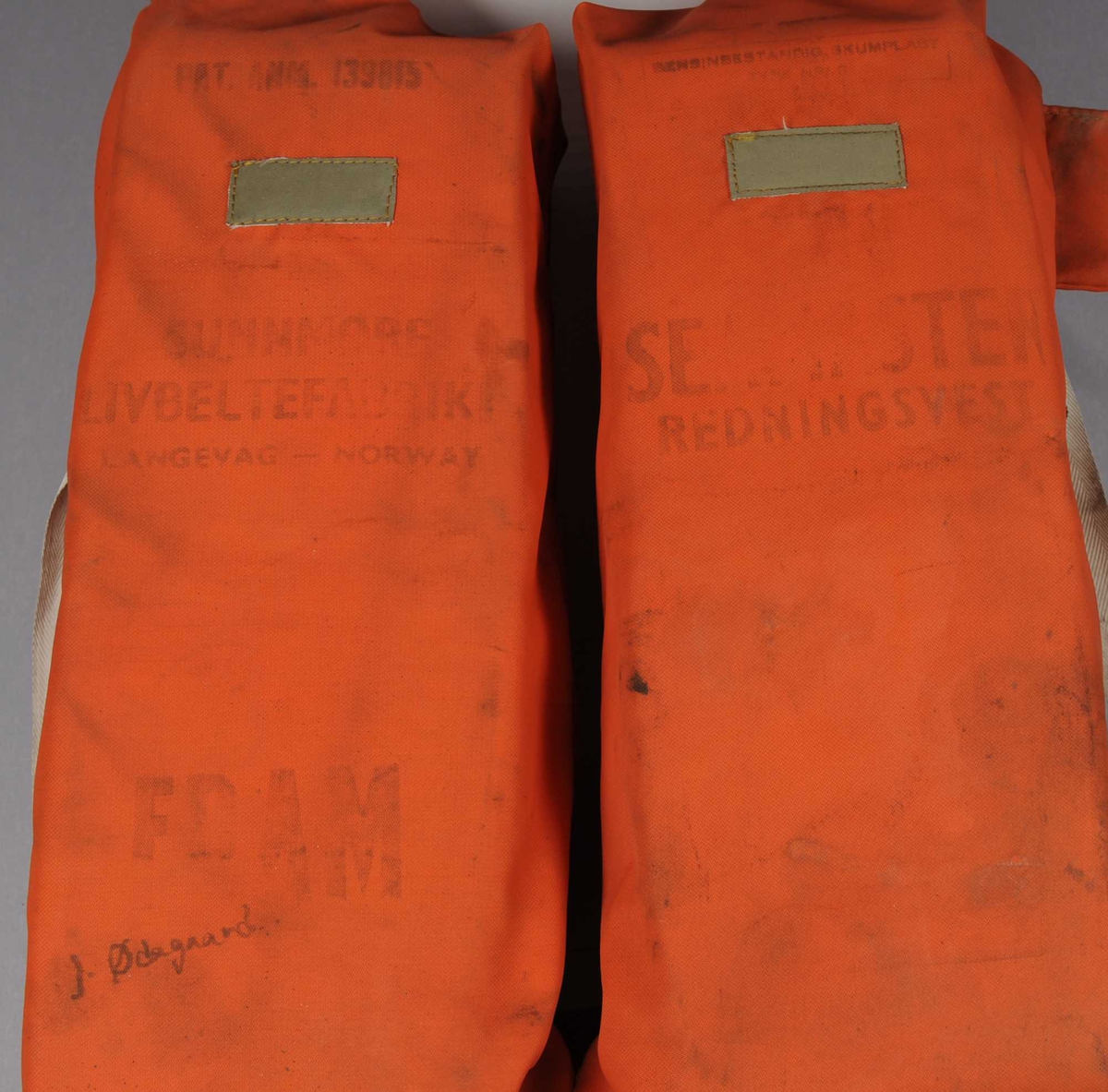 Redningsvest av orangefarget tekstil med flytemiddel inni. Det er trykt tekst og en håndskreven signatur på den ene siden. Det er remmer av tekstil.