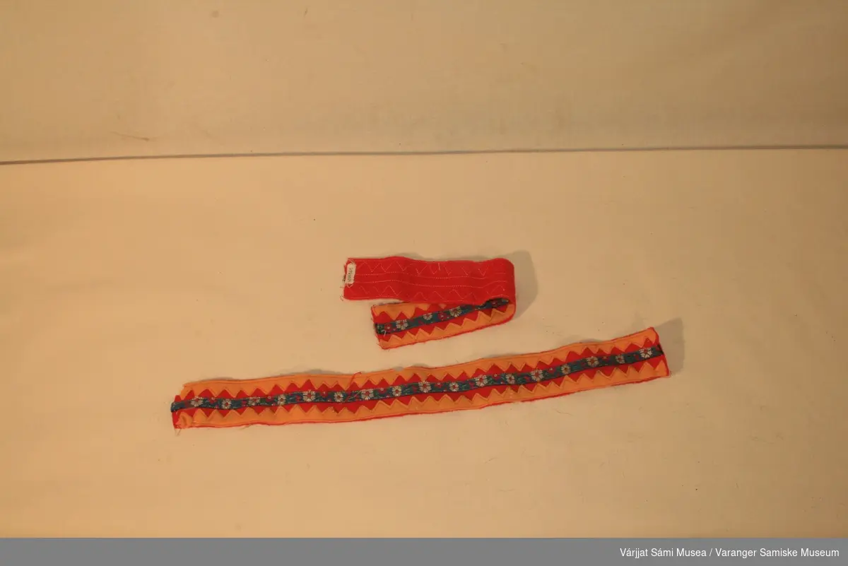 To pyntebånd. Bunnen er  rødt klede. Gule sikksakk mønster i kantene og blå mønstret bånd i midten. Pyntebåndene kan være brukt som pynt til skaller. En kan se at disse er klipt løs i fra noe.