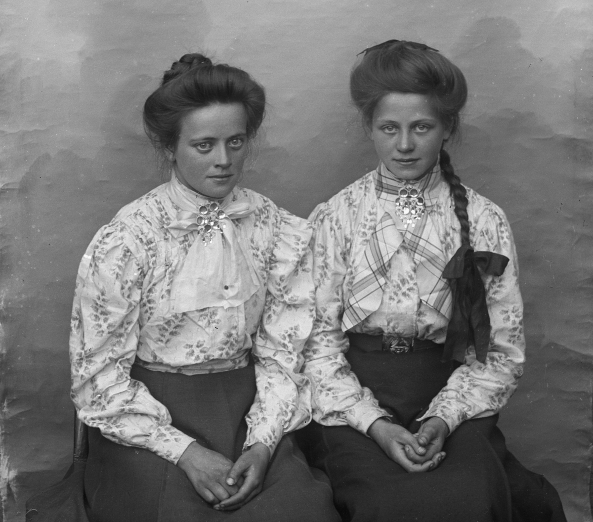 To unge jenter kledd i mønstrete lyse skjorter med søljer, den ene jenta med lang synlig flette. Lerretbakgrunn