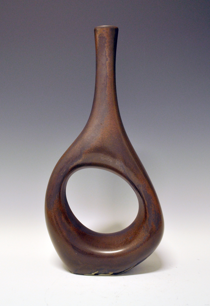Vase av porselen. Lang, smal hals, nedre del asymmetrisk form med hull i midten. Brun glasur.
Kunstner: Ellef Gryte.