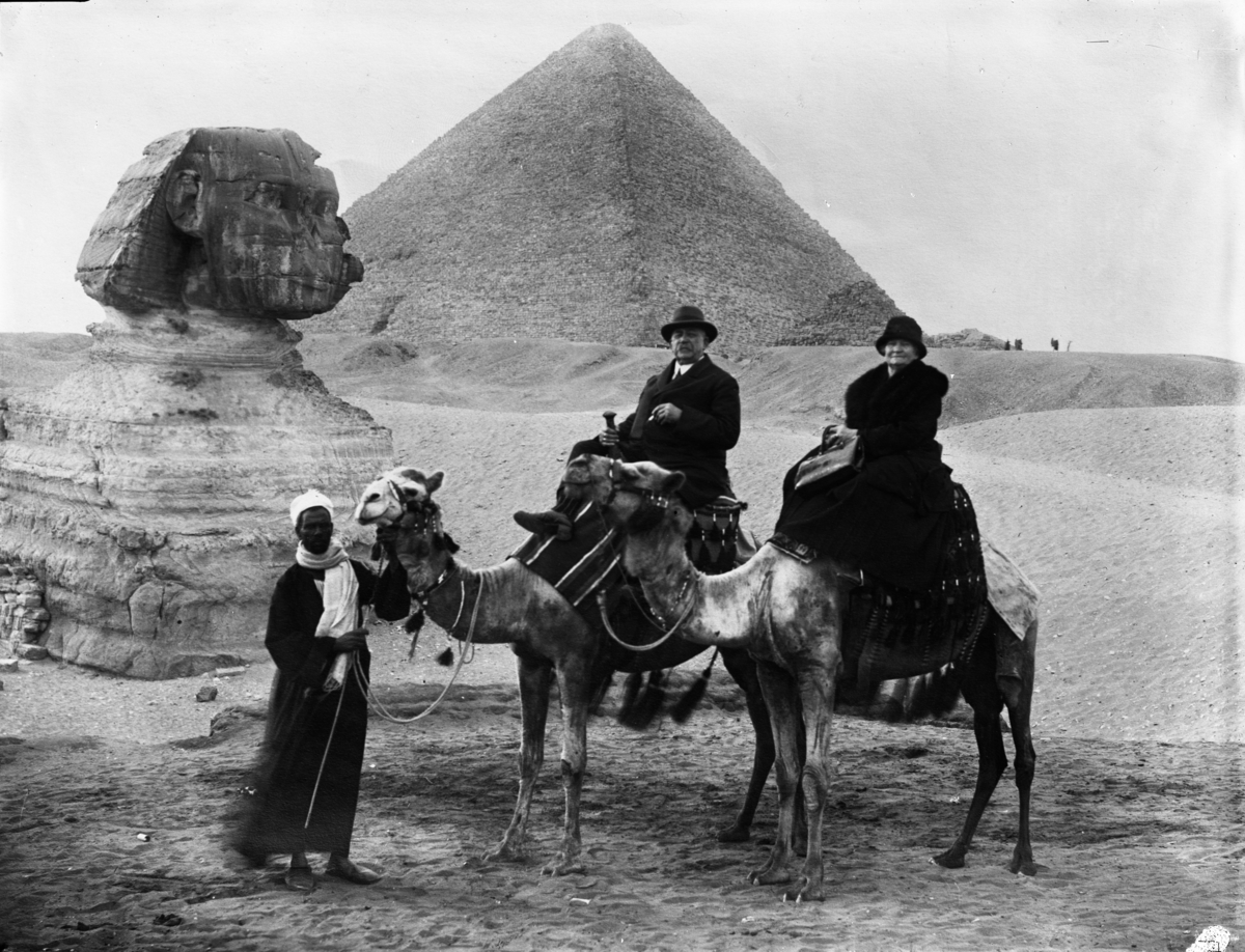 To mennesker som rir på kameler og kameleieren fotografert i Egypt med en pyramide i bakgrunnen