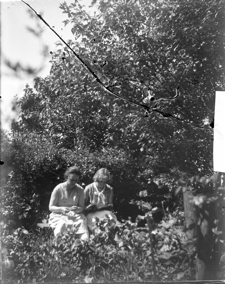 'Vy med 2 kvinnor sittande på bänk i ev. trädgård. I färd med att läsa resp. handarbeta. Text på kuvertet som plåten ligger i: ''kyrkesund 1929.'' ::  :: Ingår i serie med fotonr. 5226:1-11.'