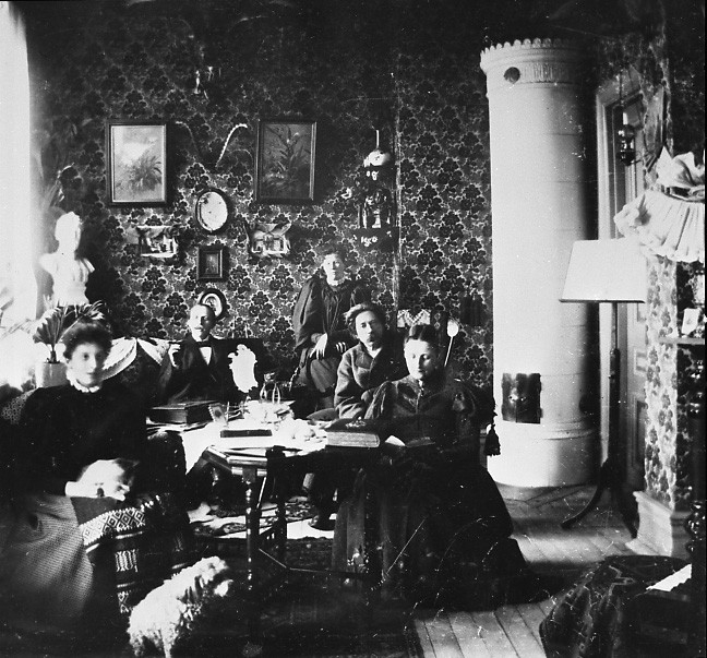 'Interiör. :: Repro av porträtt samt äldre Göteborgskarta. ::  :: 3 kvinnor samt 2 män sittande i ett möblerat rum med bl.a. en kakelugn.'