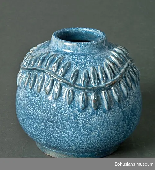 Klotrund vas med dekor av pålagd bladsinga i relief. Vit engobe med blå glasyr. I botten handskriven signatur: Greta