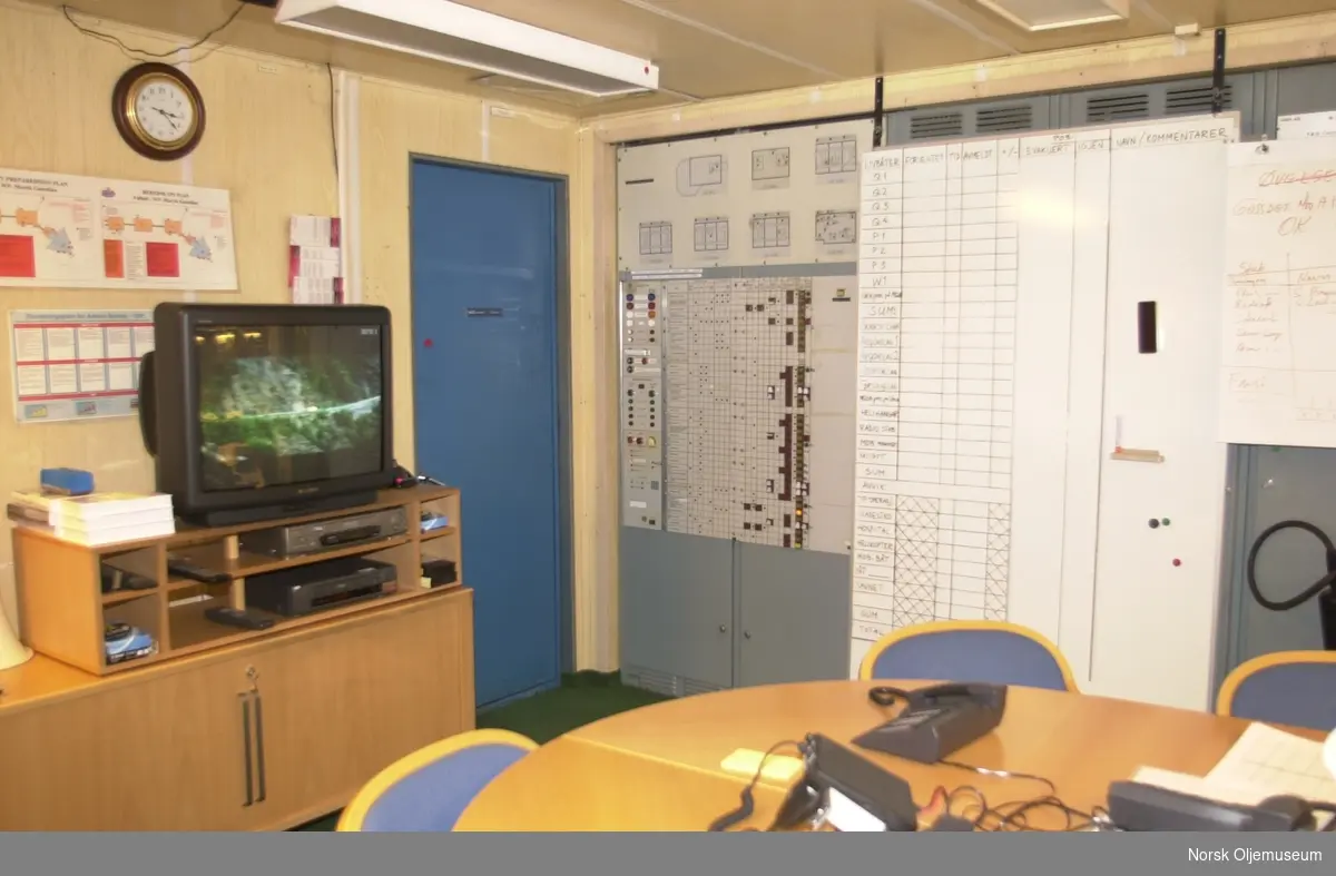 Et kontor med diverse kontrollpaneler langs den ene veggen. I forgrunnen står et rundt bord med en del telefoner og kommunikasjonsmedier. I en ende står en påskrudd tv.