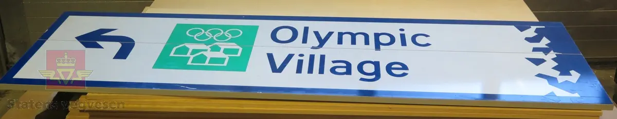 Rektangulært skilt av aluminium. Har symbol for olympisk bebyggelse, kjøreretning og teksten "Olympic Village". Flerfarget,  med grå bakside.