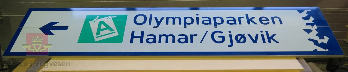 Rektangulært skilt av aluminium. Har symbol for kjøreretning og akkreditering samt teksten "Olympiaparken Hamar/Gjøvik". Flerfarget, med grå bakside.