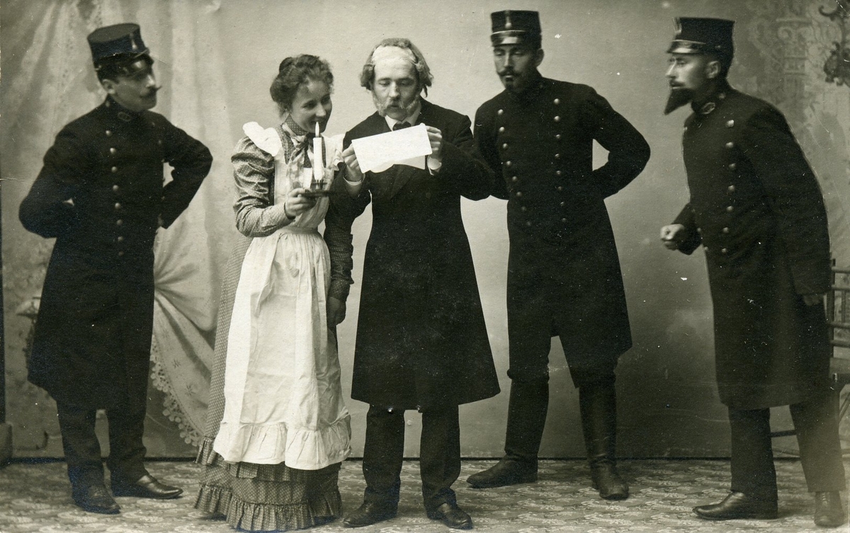 Brevkort med fotografiskt motiv: en herre som håller ett brev och en dam stående bredvid med ett ljus, båda läser brevet. Tre uniformsklädda herrar försöker nyfiket se vad som kan stå i brevet.