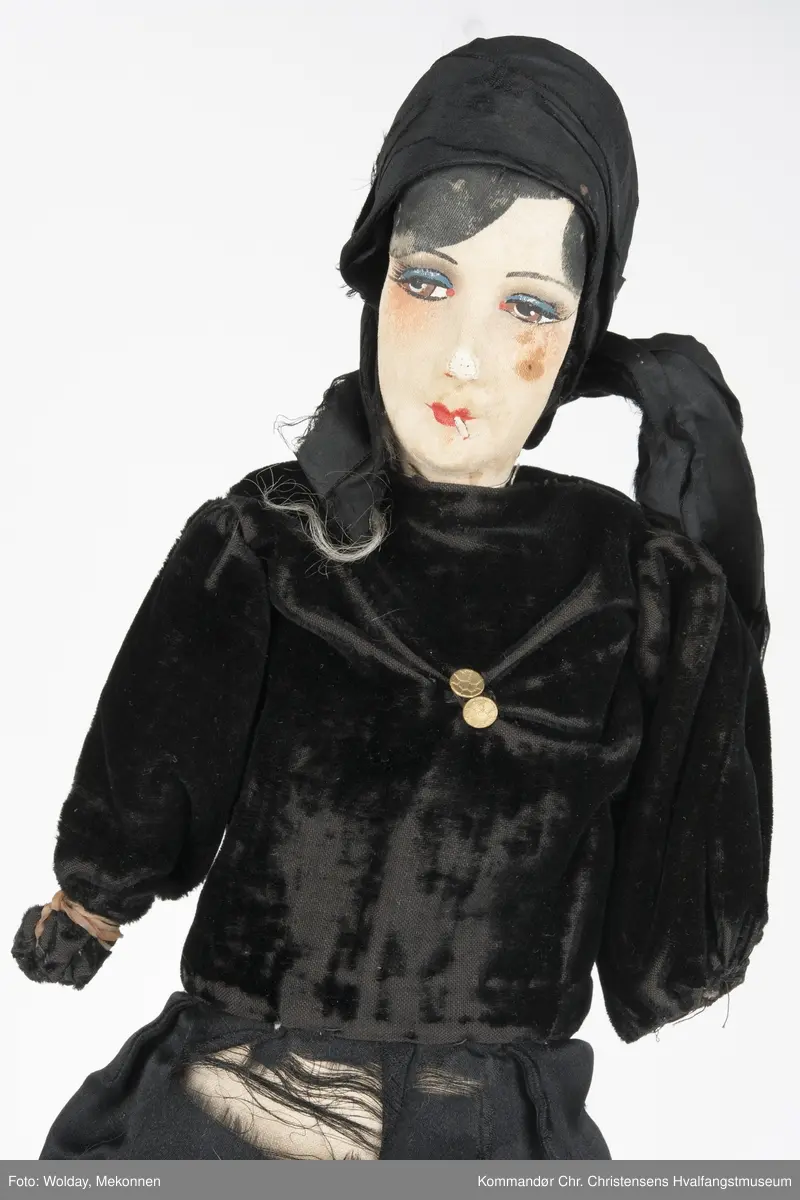 Dukken: sminket kvinneskikkelse med sigarett i munnen,  tørkle på hodet og sort todelt dress (Langermet, kort bluse, lange bukser, strømper.