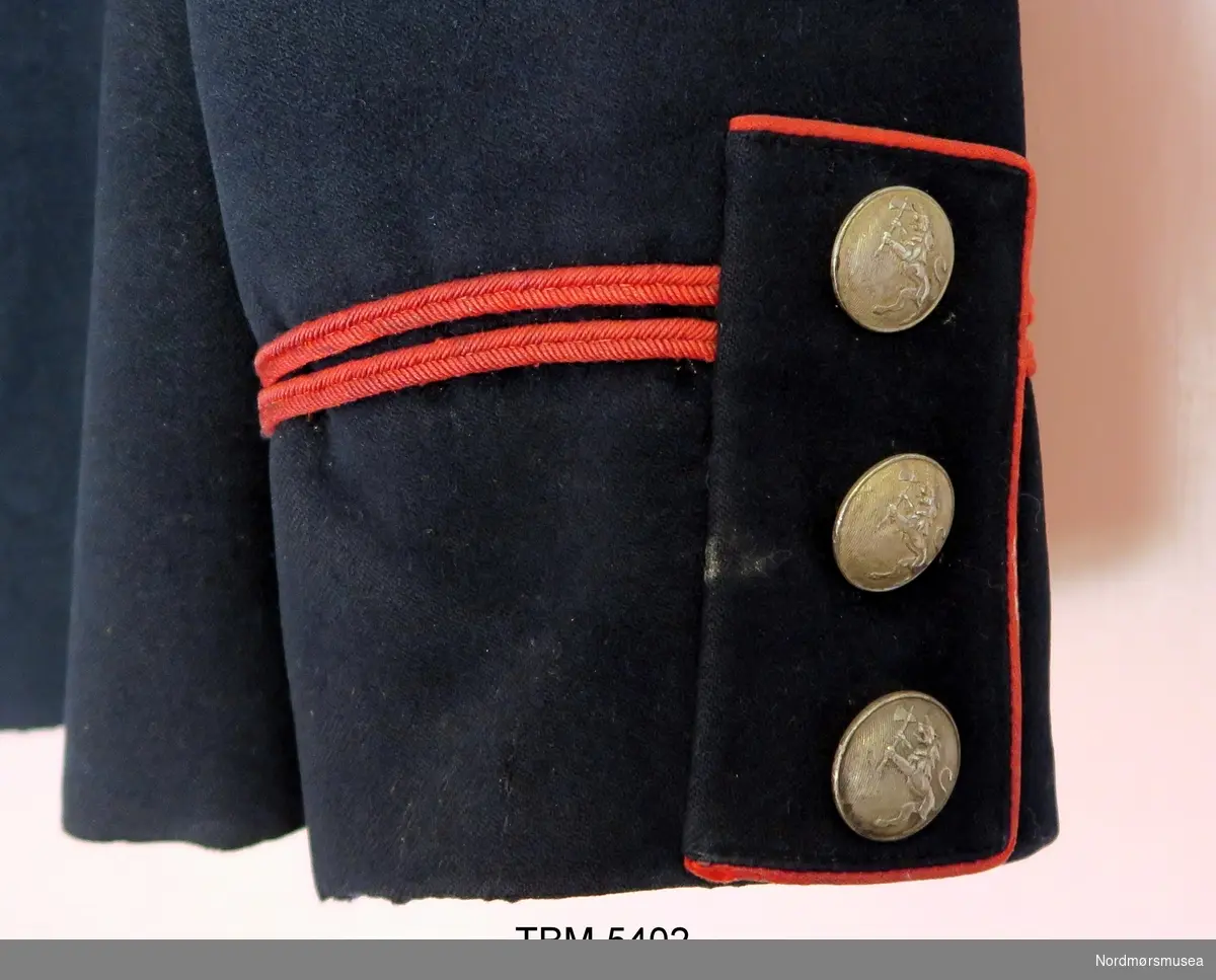 Militær jakke som er sort med raude kantar. Blanke knappar med norske løve på. På jakkekragen er det metallbokstavar, NB. Splitt med knappar og raude kantar bak.