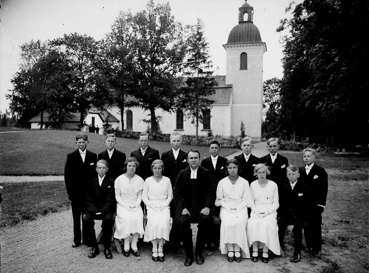 14 konfirmander, 4 flickor, 10 pojkar och kyrkoherde Areborg.
Lillkyrkas kyrka i bakgrunden.