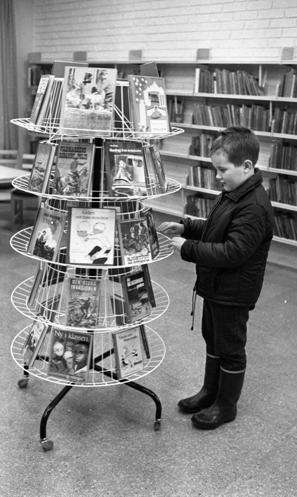 Bibliotek i Pålsboda 10 februari 1967En pojke i jacka och gummistövlar står vid en ställning med böcker och tittar på böcker.