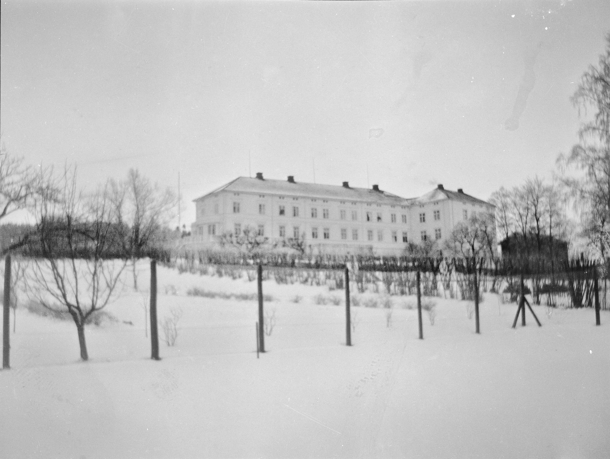 Hageanlegget i vinterskrud. En snødekkket tennisbane med hekker, busker og trær, uthus og hovedhus i bakgrunnen.