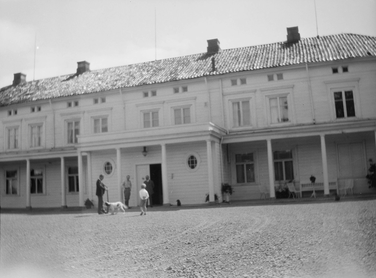 Hovedhuset på Linderud gård med gårdsplassen i forgrunnen ved sommerstid. Tre menn, en gutt samt en hund  står ved inngangsdøren. Under svalgangen står det hagemøbler og urner med blomster.