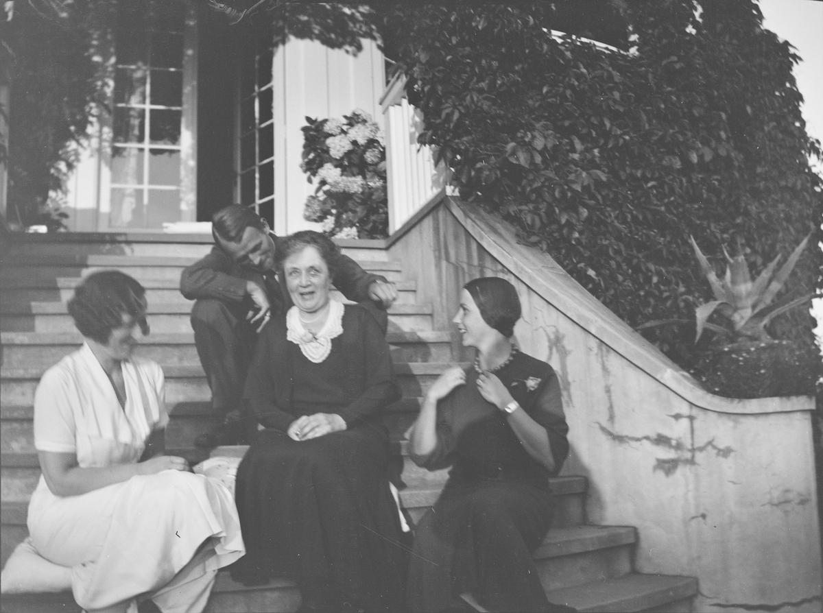 Tre kvinner og en mann sitter på trappen opp til verandaen fra den "Victorianske hagen" på Linderud Gård. De har tatt med puter som de sitter på. Verandaen er dekket av klatreplanter.