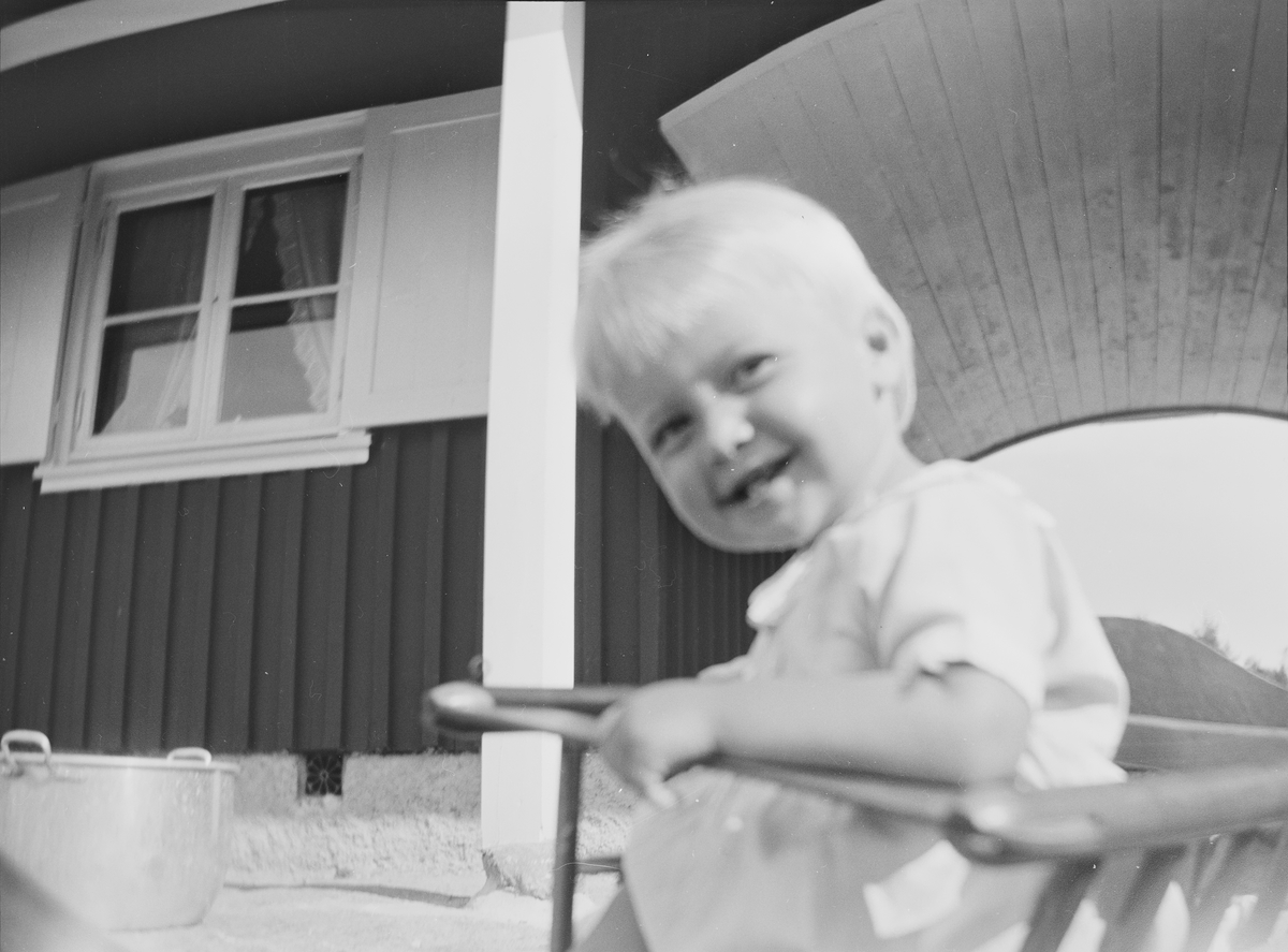 En liten gutt, "Lillefar", sitter i en barnestol, og smiler til fotografen, foran et hus som består av to deler. De to delene har felles tak, en portal utgjør skillet.