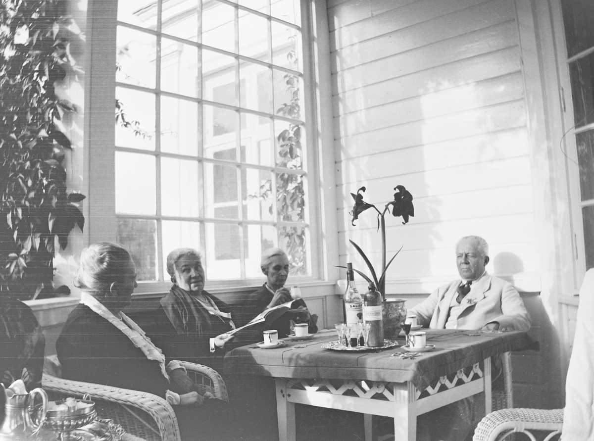 Christian Pierre Mathiesen med tre av søstrene: Julie til venstre, Louise i midten, Anna til høyre - sitter i kurvmøbler rundt et bord på verandaen på Linderud gård.