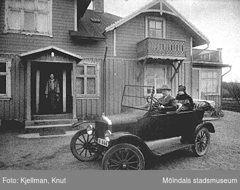 Lindome 1914-. Handlare Karl Andersson sitter i en T-Ford och hustrun sitter i baksätet. På trappan, i huset bakom bilen, står en kvinna och en hund.