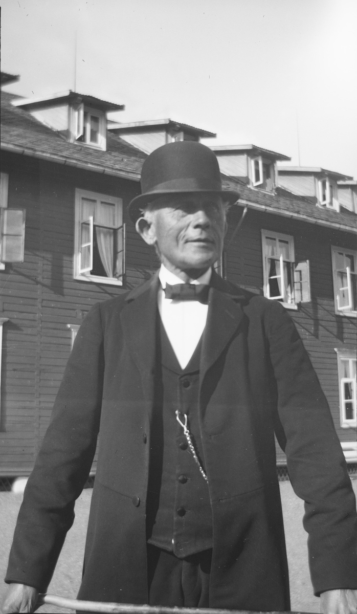 En eldre mann i dress og med bowlerhatt står utenfor en stor bygning. Han holder i en stokk eller pinne med begge hender.
