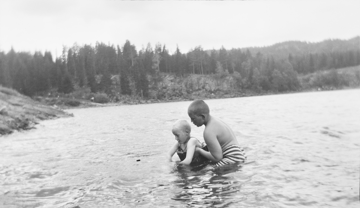 En eldre gutt hjelper en yngre gutt med svømmetrening i en innsjø på østlandet, muligens i Jeppedalen. Den yngste gutten har på svømmebelte.