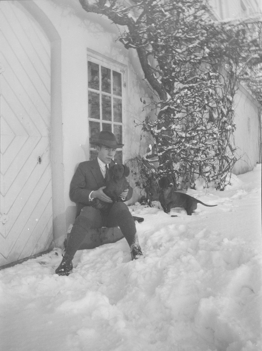 Iacob Ihlen Mathiesen sitter, på en stein eller lignende, utenfor en dør i grunnmuren til hovedhuset på Linderud Gård. Han har med seg to dachser.