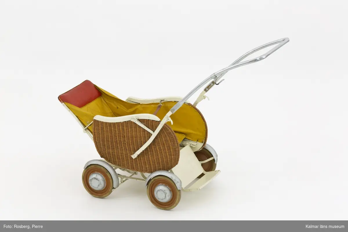 KLM 28596:2. Barnvagn, sittvagn, metall, trä, färg, textil, läder. Sittvagn för barn, ihopfällbar med stomme av järn och fyra hjul och reglerbart fotstöd av trä och järn. Sittkorgen har fastspikad klädsel av textil, målad med gul färg, och rödmålat, stoppat nackstöd. Ryggstödet är reglerbart i två lägen. Tvärs över sitsen hänger ett uppspänt läderband som fästs med hyska för barnets säkerhet. Ihopfällbart och reglerbart handtag av järn, övre delen kromat, och en krok på vänster sida. Vagnens sidor har ett korgflätat yttre lager, vitmålade sargar och silvermålade stänkskärmar och navkapslar.