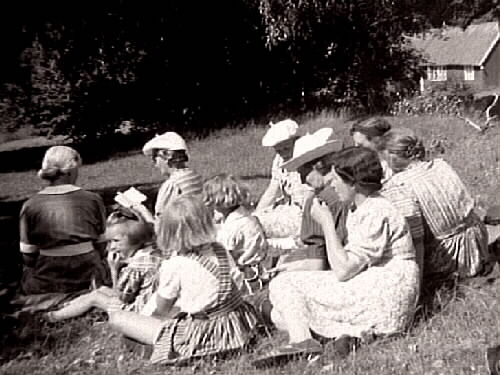 Utflykt. En grupp kvinnor och några barn sitter på en äng. En av kvinnorna längtfram håller upp och läser i en liten bok. I bakgrunden syns en skogsdunge och ett hus.