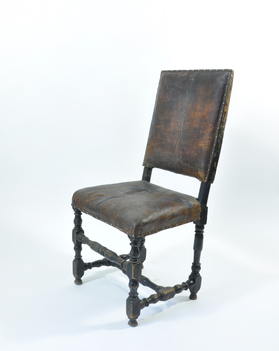 Høyrygget stol trukket med mørkbrunt lær, festet med metallnagler (messing). Beina og bindingsbrettene er dreid. Beinkonstruksjonen er svartmalt.