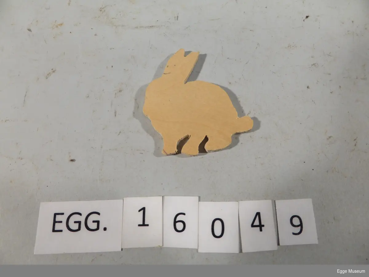 En liten kanin som er skåret ut av kryssfinèr. Fargelagt på ene siden. Knainn er grå og spiser en oransje gulerot.