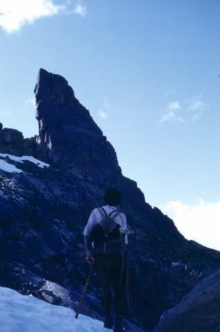 Mann med ryggsekk og tau på vei opp mot smal spiss fjellformasjon. Fjellklatring. hvor
