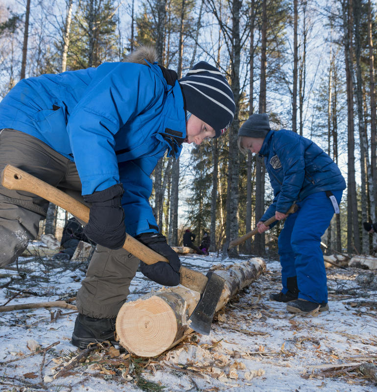 Bildet viser to gutter som fjerner bark og kvister med øks på en stokk på bakken.