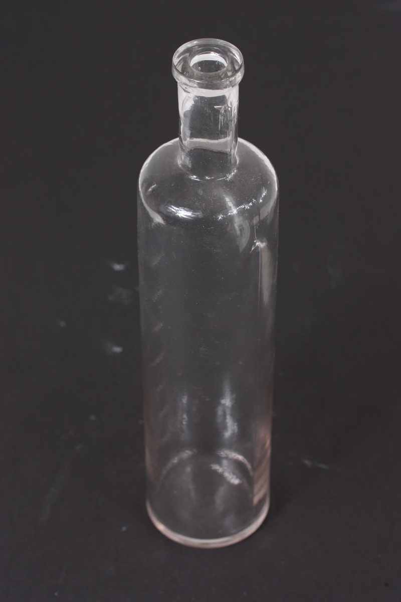 Slank sylinderisk flaske som kragar inn og endar i ein smal lang tut. Innstløypte strekmål på flaska.