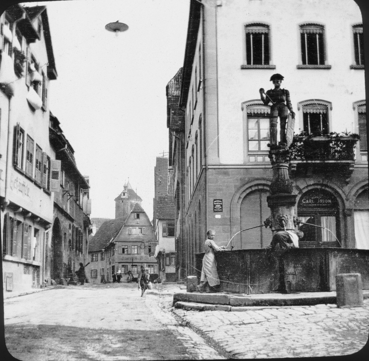 Skioptikonbild med motiv av fontän "Jörgle" på gata i Besigheim.
Bilden har förvarats i kartong märkt: Resan 1907. Besigheim 8. 29. Text på bild: "Neckar Eng."
