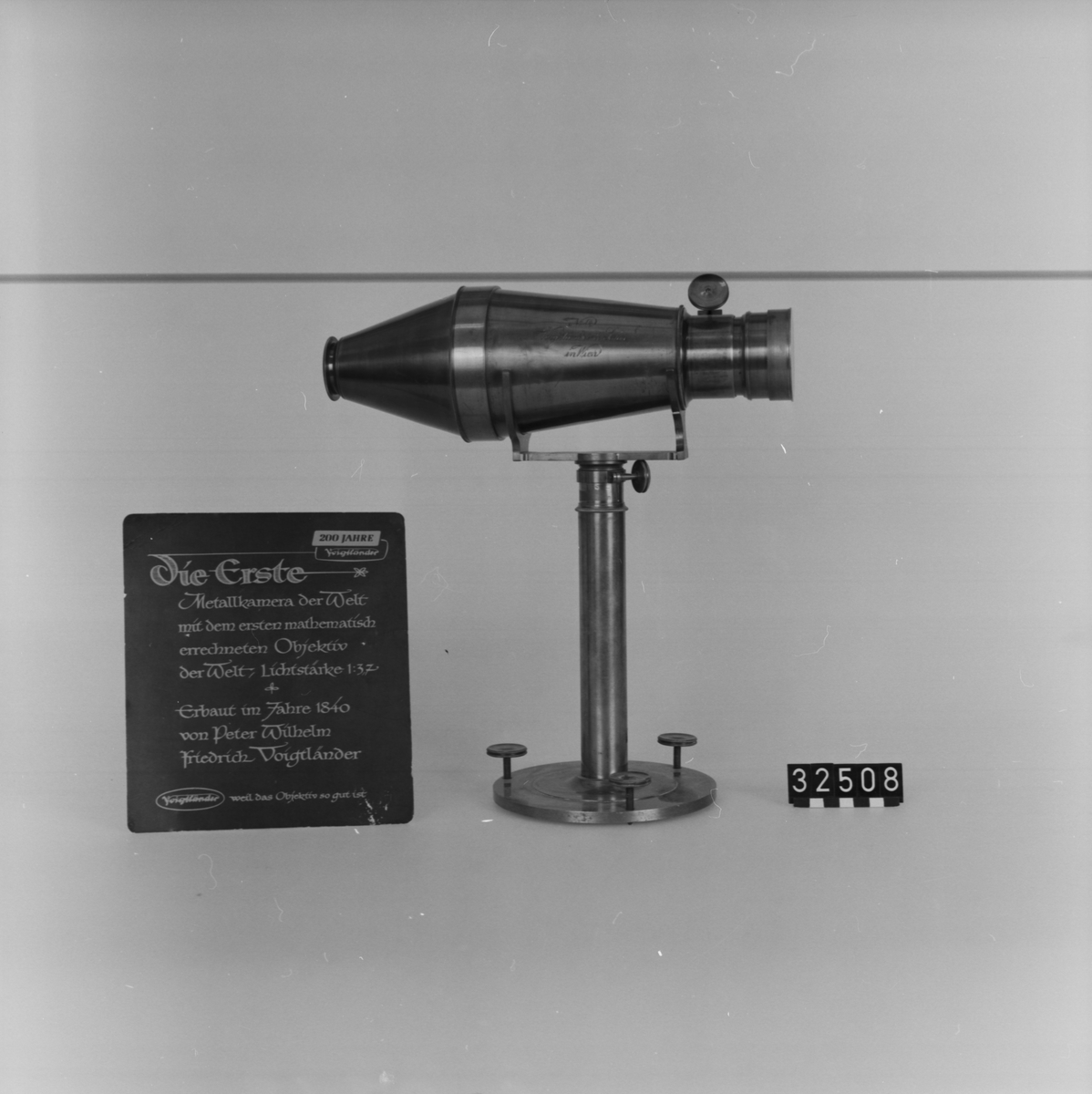 Kopia av Voigtländers första metallkamera för Daguerreotyper (mässingstub). Originalet tillverkades på 1840-talet. Denna kopia med nr. 84 tillverkades troligen c.a. 1930. Ljustyrka 1:3,7. Bildstorlek c.a. 100 mm/diam. Kamerakropp på fot med ställskruvar, två byglar för uppläggning av kameran. Stativ, diameter: 155 mm, höjd: 295 mm.