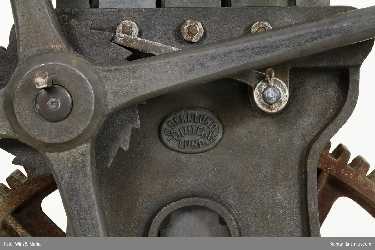 KLM 21763. Garnpress. Användes för pressning och buntning av färgat garn. Av järn. På ena sidan av stommen är gjuterifirmans namn präglat: C. BERNLUNDS GJUTERI LUND. Sidorna består av en stomme med fyra fötter. På ena sidan ett handtag, hjul, som vid vridning driver ett större kugghjul. På kugghjulets axel sitter två armar som pressar en järnplatta uppåt. Ena armen har gått av. På järnplattan placeras garnet och pressas upp mot taket som består av breda järnstänger som kan fällas upp och ned. Vid buntning lades även ett snöre om garnet och när garnbuntarna var hoppressade knöts dessa samman.