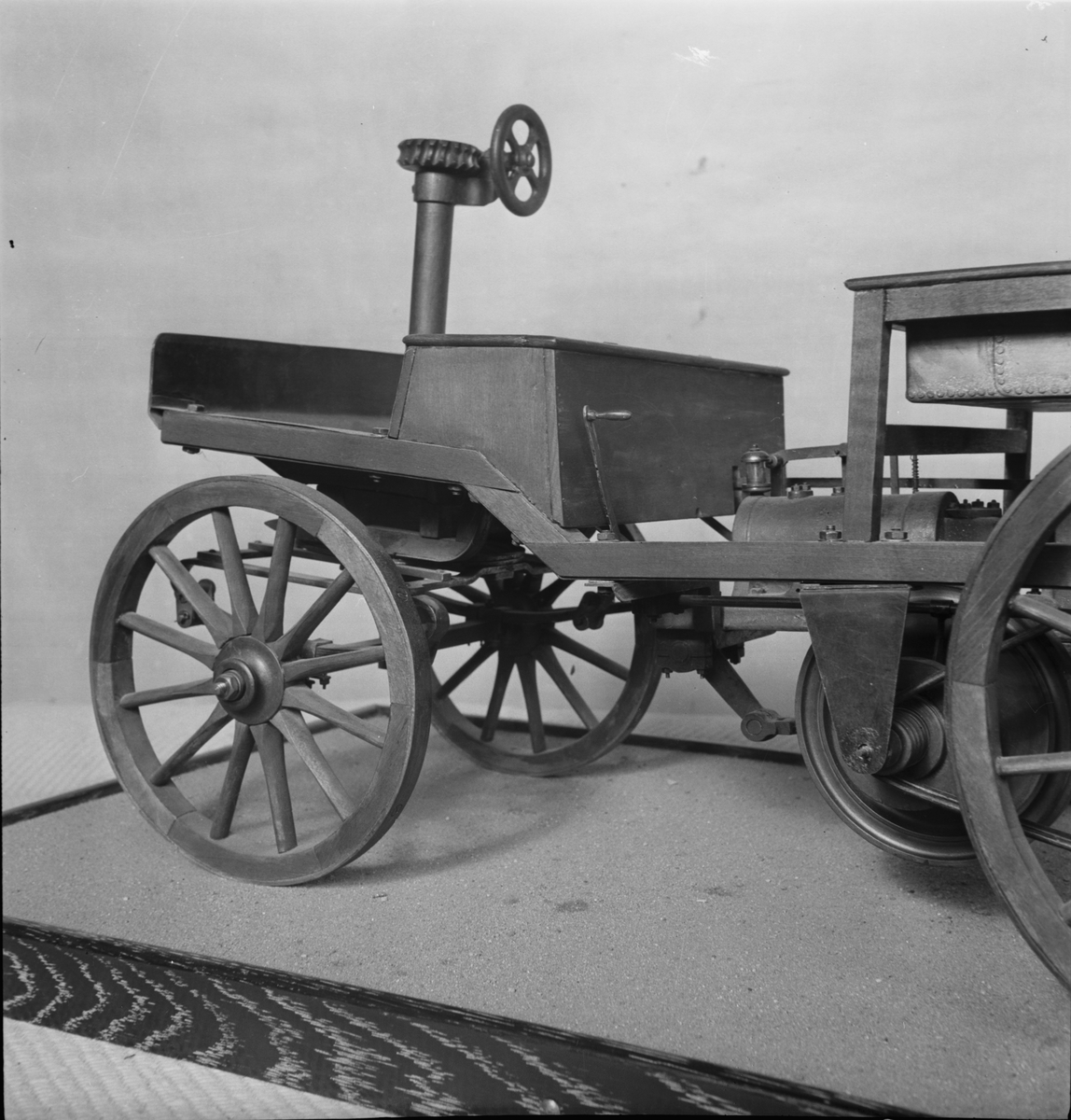 Modell i skala 1:5 av bensinmotordriven bil med hästvagnsliknande kaross. Motorn tillverkad av Märky, Bromovsky & Schultz 1888. Kraftöverföring från motorn med remmar till remskiva på bakaxeln. Ram av trä, framvagn med halvelliptiska fjädrar. Trähjul skodda med järnband. Förebilden har 2 m axelavstånd. Monterad på en platta 700 x 400 mm  Encylindrig luftkyld fyrtaktsmotor Effekt cirka 4 hk